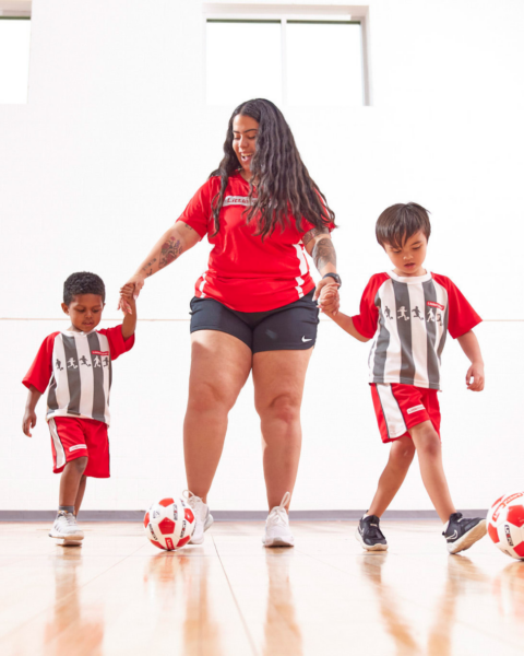 A coach stood up holding two children's hands while they attempt to kick Little Kickers footballs across a sports hall. 